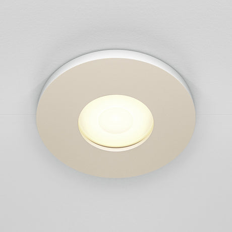 The Stark Fixed Round Recessed Downlight - White, featuring a glowing GU10 bulb, is set in a smooth surface. Its modern design emits soft, warm light and boasts an IP65 rating for dependable use in moist areas.
