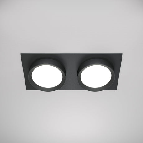 The Hoop Twin Fixed Rectangular Recessed Downlight in black elegantly features a sleek rectangular base holding two round white LED downlights, casting a soft glow against the plain white ceiling.