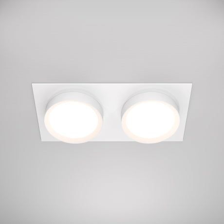 The Hoop Twin Fixed Rectangular Recessed Downlight in white, featuring dual LED circular lights and a rectangular base, provides a soft glow. Its modern, minimalistic design contrasts beautifully against the plain gray ceiling.