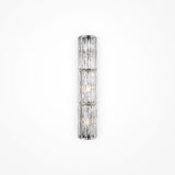 The Facet 3 Light Long Wall Light in chrome is a vertical cylindrical glass sconce with crystal and faceted panel detailing, emitting a soft, diffused light. Mounted on a plain white wall, it adds an elegant and modern touch to any space.