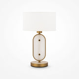 The Marmo Table Lamp in gold features a white cylindrical lampshade, a stylish base with a gold frame surrounding an oval natural stone panel, and rests on a circular gold platform.