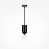 The Memory Hourglass Pendant Light features a black cylindrical fixture with a white frosted glass shade, hanging elegantly from a thin black wire and round ceiling mount, making it an ideal addition to modern interiors for ambient lighting against plain white backdrops.