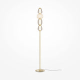 The Pattern LED Floor Lamp - Gold is tall and modern with a slim gold base and pole, featuring four stacked textured glass orbs in clear and frosted finishes. Its black power cord enhances energy efficiency against a plain white backdrop.