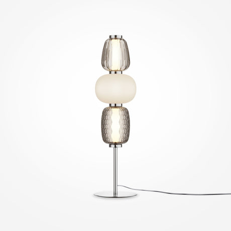The Pattern LED Table Lamp - Chrome offers a modern look with its chrome finish and metallic base, featuring three stacked glass shades: textured clear on top, smooth white in the middle, and textured amber at the bottom. A black power cord enhances its contemporary appeal.
