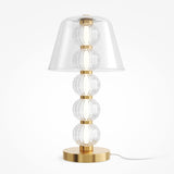 The Amulet LED Table Lamp features a gold brass base with five ribbed clear glass spheres, a wide transparent conical shade, and a white power cord.