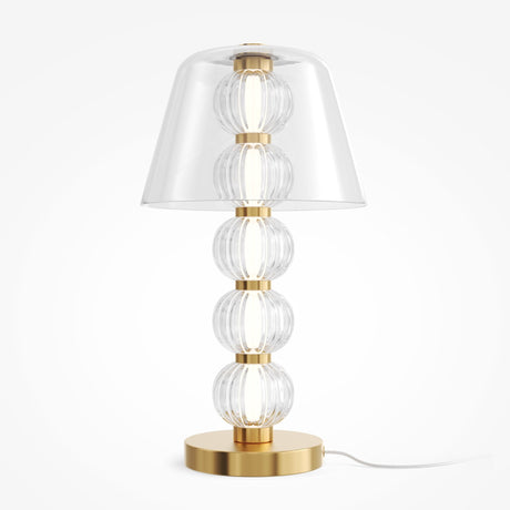 The Amulet LED Table Lamp features a gold brass base with five ribbed clear glass spheres, a wide transparent conical shade, and a white power cord.