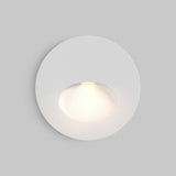The Bil Outdoor LED Recessed Wall Light - White is a modern, circular fixture featuring energy-efficient LED technology. Its minimalist design emits a soft glow from the recessed center, creating a halo effect on the surrounding plain gray surface.