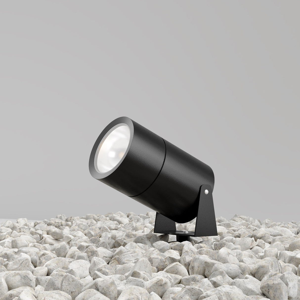 The Bern Outdoor LED Spike Light 15W 3000K in graphite, rated IP65, is mounted on a stand amid white pebbles against a plain light gray wall. Though off, its design suggests outdoor versatility.