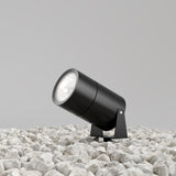 The Bern Outdoor LED Spike Light 15W 3000K in graphite, rated IP65, is mounted on a stand amid white pebbles against a plain light gray wall. Though off, its design suggests outdoor versatility.