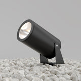 The Bern Outdoor LED Spike Light 30W 3000K in graphite, with a cylindrical design and tilted stand, is set on white pebbles. This IP65-rated light shines upwards against a gray backdrop, perfect for outdoor conditions.