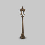 A vintage-style street lamp, Fleur 1 Light Pillar Lantern in black, with intricate detailing and a clear PULEGOSO glass lantern, stands against a plain gray background.