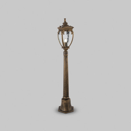 A vintage-style street lamp, Fleur 1 Light Pillar Lantern in black, with intricate detailing and a clear PULEGOSO glass lantern, stands against a plain gray background.
