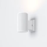 The Rando Outdoor Wall Light - White is a minimalist, wall-mounted cylindrical lamp with vertical grooves, emitting soft light. Designed for architectural lighting, it boasts an IP54 rating and attaches to a white rectangular base on the wall.