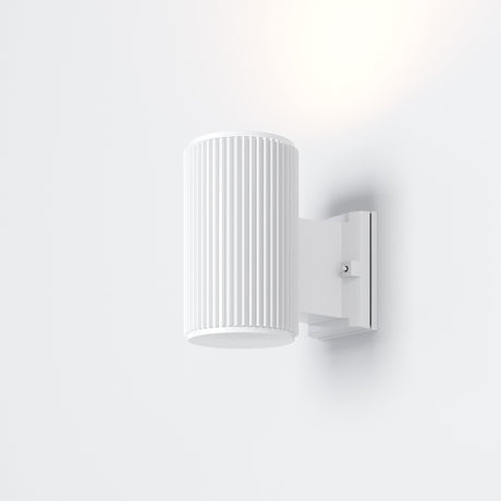The Rando Outdoor Wall Light - White is a minimalist, wall-mounted cylindrical lamp with vertical grooves, emitting soft light. Designed for architectural lighting, it boasts an IP54 rating and attaches to a white rectangular base on the wall.