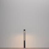 The Mill LED Short Bollard Light - Graphite, with its minimalist cylindrical black base and narrow vertical light tube, elegantly stands on a plain surface. Its graphite finish enhances the soft glow on the neutral gray wall behind it.