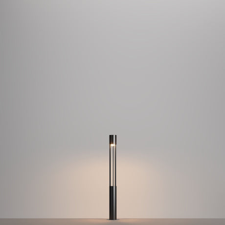 The Mill LED Short Bollard Light - Graphite, with its minimalist cylindrical black base and narrow vertical light tube, elegantly stands on a plain surface. Its graphite finish enhances the soft glow on the neutral gray wall behind it.
