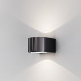 The Blow Outdoor LED Up and Down Wall Light in graphite is installed on a white wall. Rated IP54, this modern fixture provides soft white illumination both upwards and downwards, achieving an elegant minimalist effect.