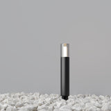The Glide Outdoor Long Spike Light - Graphite, a contemporary black lamp with a transparent top, is illuminated and stands on white pebbles against a gray background.