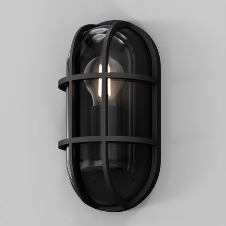 The Candle Outdoor Wall Light in graphite features an industrial-style design with a metal grid and clear glass cover, showcasing the light bulb. This oval fixture, mounted on a plain gray wall, offers a modern industrial look with IP65-rated durability.