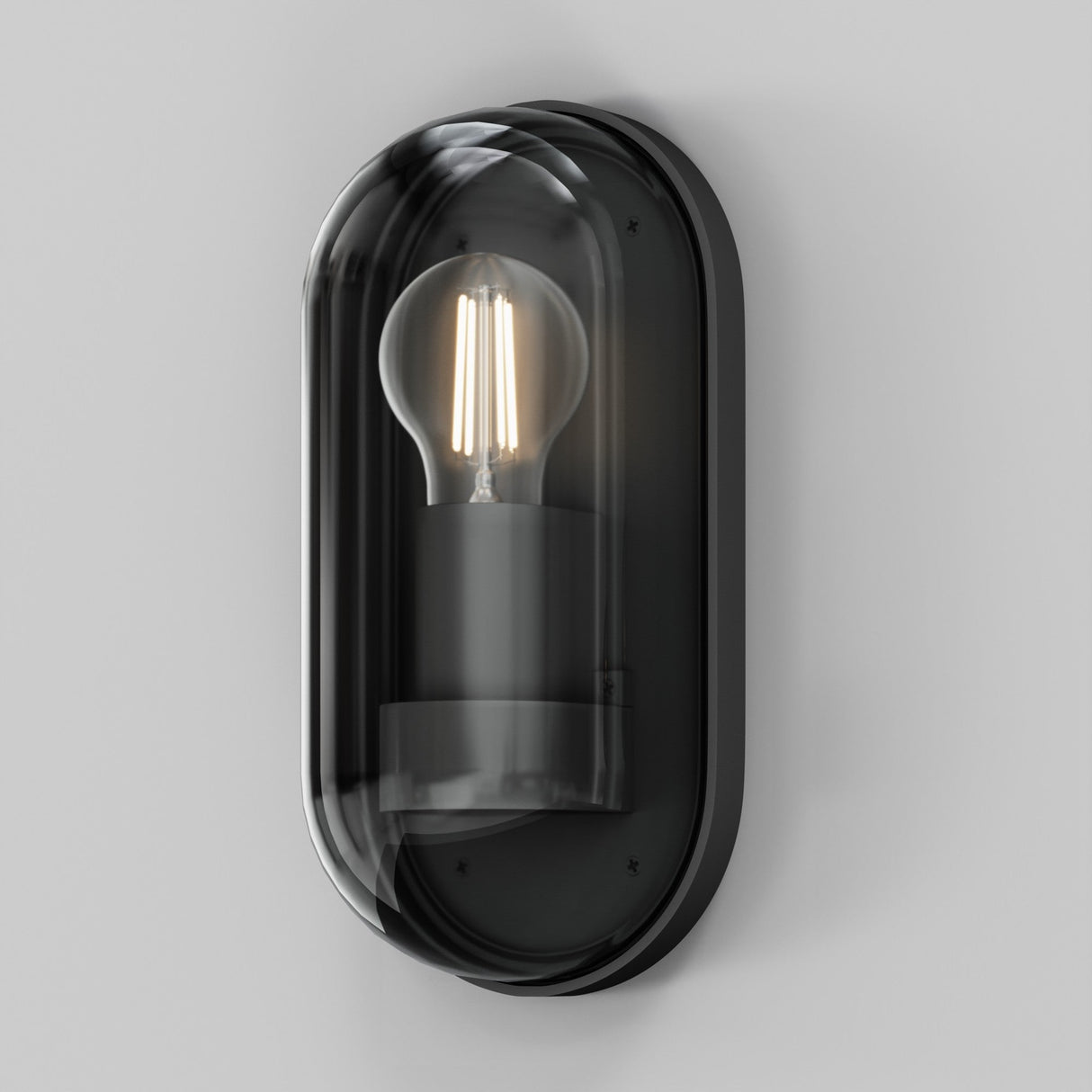 The Candle Outdoor Wall Light - Graphite is a modern wall sconce with a minimalist black frame, clear glass cover, and visible filament bulb that casts a warm glow. Its oval design brings contemporary loft-style charm to any space against a plain white background.