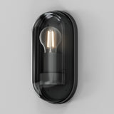 The Candle Outdoor Wall Light - Graphite is a modern wall sconce with a minimalist black frame, clear glass cover, and visible filament bulb that casts a warm glow. Its oval design brings contemporary loft-style charm to any space against a plain white background.
