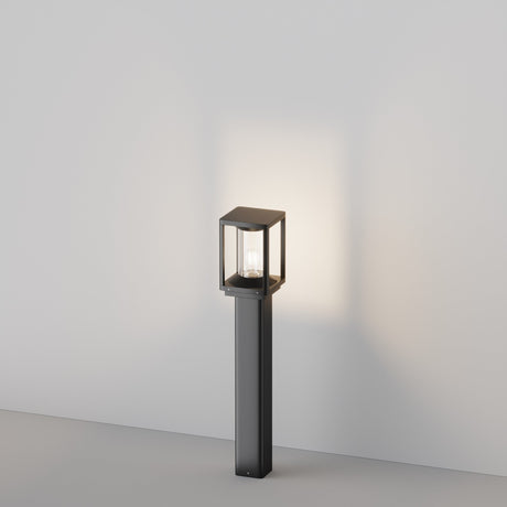 The Cell Bollard Light 75cm - Graphite enhances your outdoor space with its sleek graphite frame and clear glass panels. Emitting a warm glow against a light gray background, its IP54 rating guarantees durability while casting gentle shadows for an inviting ambiance.