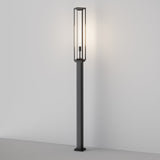 The Cell Bollard Light 220cm - Graphite features a tall, slim frame with a rectangular glass enclosure for soft ambient lighting. Placed against a stark white wall on a light surface, it perfectly captures modern minimalist design akin to sleek outdoor lighting.