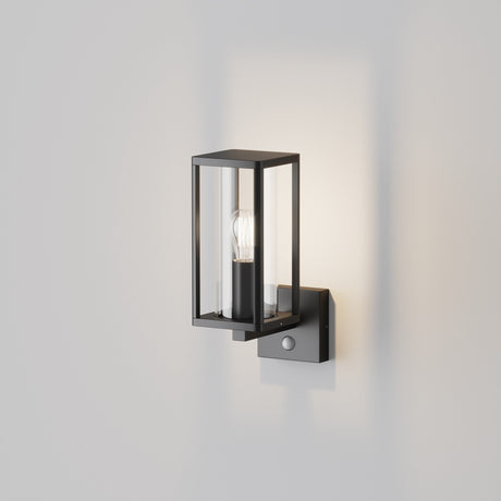 The Cell Outdoor Motion Sensor Wall Light - Graphite showcases a lantern-style design with a black metal frame around an exposed bulb. Mounted on a square base against a white wall, it emits an inviting glow and is IP65-rated for reliable outdoor use.