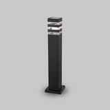 The Unter den Linden Bollard Light - Black is a modern, rectangular outdoor light featuring a sleek black base and a transparent top section for illumination. Built with corrosion-resistant materials for durability, it showcases horizontal lines against a plain gray background.