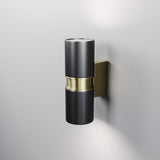 The Bowery Outdoor 2 Light Wall Light - Black (O582WL-02B) with brass accents and a sleek cylindrical design enhances a light gray wall. Its modern black and gold look, with upward and downward lighting, offers a soft glow. Ideal as an outdoor fixture with its durable IP54 rating.