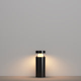 A minimalist black table lamp with a cylindrical base and flat top rests on a grey surface. Its energy-efficient design mirrors the Olten Bollard Light 30cm in Graphite, emitting a warm glow against the grey background for a calm, cozy ambiance.