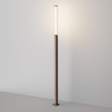 The Lit LED Tall Bollard Light - Brown is a modern, slim floor lamp with energy-efficient lighting and a cylindrical top. Its minimalist design casts a soft glow against white walls, featuring a sleek brown base and pole for a contemporary look.