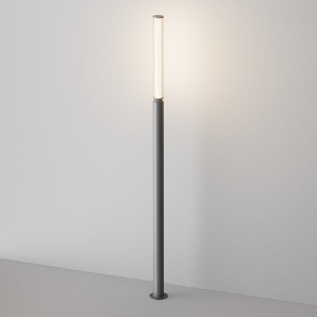 The Lit LED Tall Bollard Light - Graphite is a modern floor lamp with an energy-efficient system, featuring a tall cylindrical pole and slim vertical light source emitting a warm glow, standing against a plain white wall on a light gray floor.