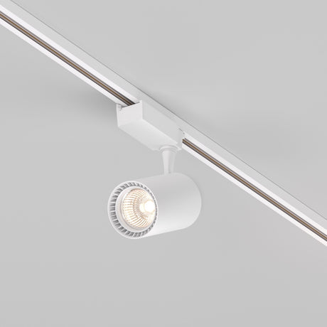 The Vuoro S LED Single Phase Unity Track Spotlight 26W 3000K in white is mounted on a ceiling rail and angled downward. Its lit bulb casts a warm glow, while the simple cylindrical design and adjustable swivel make it ideal for Scandinavian-inspired interiors.