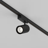 The Vuoro S 3-Circuit Trinity Track LED Spotlight 26W 4000K in black, with its cylindrical fixture and aluminum finish, is ceiling-mounted on a track, providing a sleek look and soft, focused glow to enhance the plain white ceiling for versatile lighting solutions.