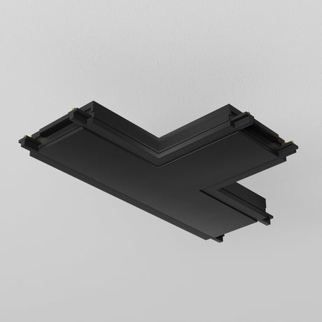 Mounted on a white wall, the black geometric shape with sharp edges resembles the Radity Magnetic Track T-Shaped Power Connector, featuring sleek, layered profiles and small golden connectors at the corners.