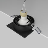 Close-up of an Atom Single Tilt Square Recessed Downlight in black, showcasing metal springs for installation. The fixture is partially placed in a ceiling cutout with hanging wires and features an adjustable tilt mechanism for directional lighting, but lacks a bulb.