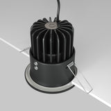 The Zoom LED Dimmable Round Recessed Downlight, 12W 4000K in black, is partially installed in a white ceiling. It includes vertical heat-dissipation fins, two mounting clips, and a top-attached cable. Its IP65 rating ensures durability while emitting crisp 4000K light.