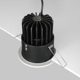 The Zoom LED Round Recessed Downlight 12W in white is partially embedded in a ceiling, featuring a cylindrical body with vertical grooves and adjustable clips. It offers high color accuracy with a CRI 90 rating, while a cable extends from the top of the fixture.
