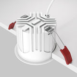 The Alfa LED Dimmable Fixed Round Recessed Downlight 15W 3000K - White blends elegance and stability, featuring a white circular design with a geometric top. Its attached to a white surface edge using two metal spring clips with red handles, resembling an LED downlight.