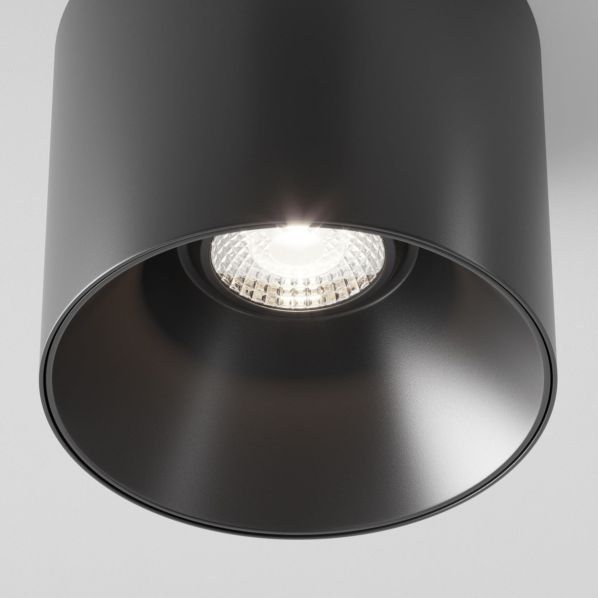 Close-up of an Alfa LED Dimmable Fixed Round Surface Downlight 15W 4000K in black, featuring a glowing bulb that casts cool white light downward; the fixture boasts a smooth cylindrical design and is mounted on a white ceiling.