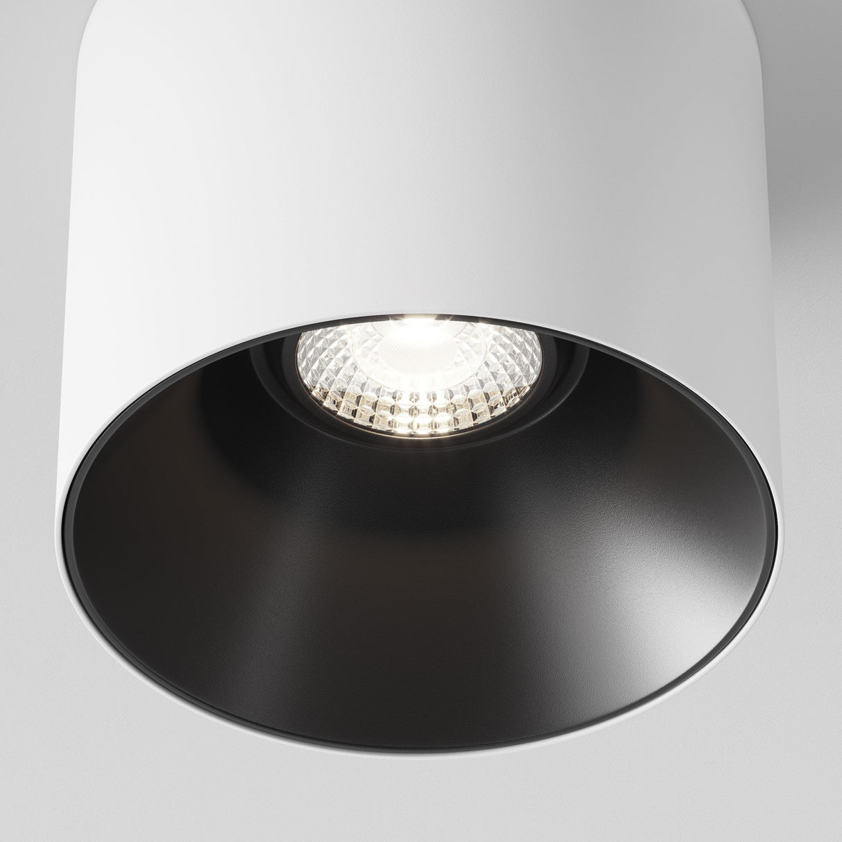 A close-up of the Alfa LED Fixed Round Surface Downlight 12W 4000K shows a fixture with a cylindrical white casing and a dark interior. The illuminated cool white bulb emits a bright glow, enhancing the modern aesthetic against the plain light-colored ceiling.
