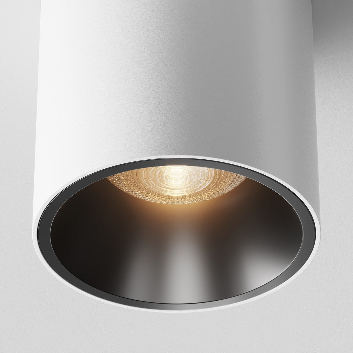 Close-up of the Alfa LED Round Surface Downlight 12W 3000K in white and black. The warm white LED light casts a cozy glow from its black-lined cylinder, perfect for modern interiors, with the textured bulb subtly visible against the plain white ceiling.