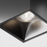 A close-up of a modern recessed ceiling light fixture featuring the Alfa LED Square Surface Downlight 12W 4000K in black. Its design includes a reflective surface and circular bulb emitting cool white light, set against a pristine white ceiling.