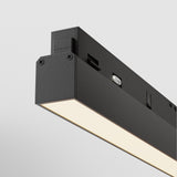 A close-up of the Basis LED Linear Track Light for Magnetic Exility, showcasing its modern rectangular design with a black casing. The illuminated 24W 3000K light casts a soft glow, embodying minimalistic elements ideal for contemporary interiors against a white background.