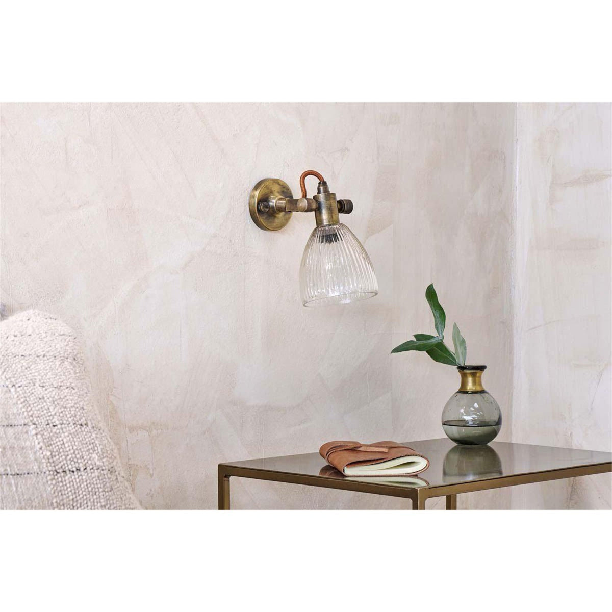 Rarni Recycled Glass Wall Light in antique brass with a ribbed clear glass tulip shade, mounted above a side table in a living room setting.