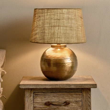 Rasan Table Lamp Base in antique brass, a spherical iron base with visible welded seams, paired with a woven textured shade on a wooden bedside drawer unit.