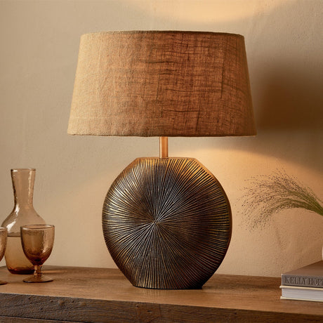 Ramvas Etched Table Lamp Base in antique brass with a sunburst-etched disc base and tan linen drum shade, styled on a wooden sideboard beside amber glassware.