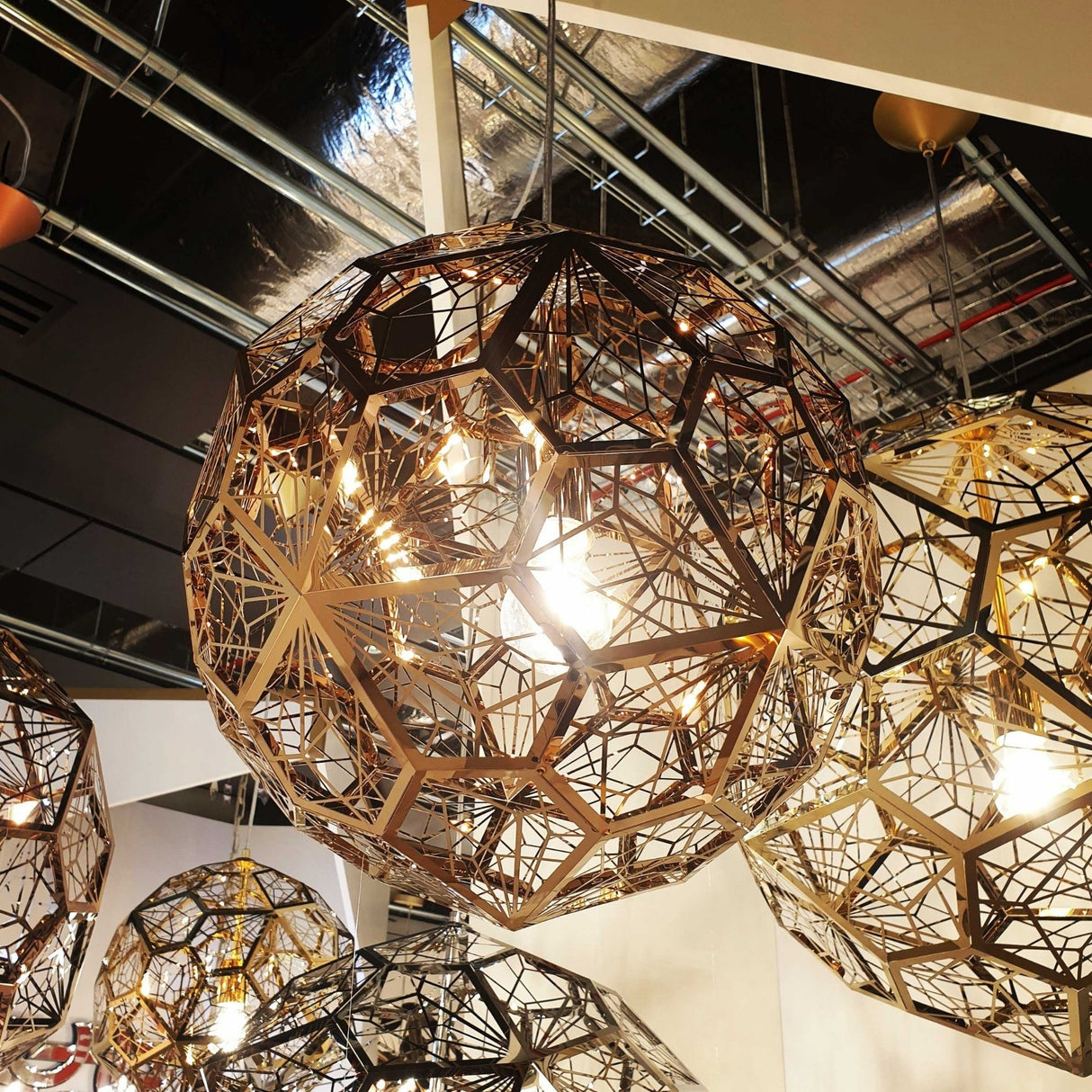 The Rango Acid Large Pendant Light in copper features an intricate wireframe design with exposed bulbs, hanging from the ceiling. Industrial pipes and ducts in the background enhance its striking industrial lighting aesthetic.