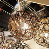 The Rango Acid Large Pendant Light in copper features an intricate wireframe design with exposed bulbs, hanging from the ceiling. Industrial pipes and ducts in the background enhance its striking industrial lighting aesthetic.